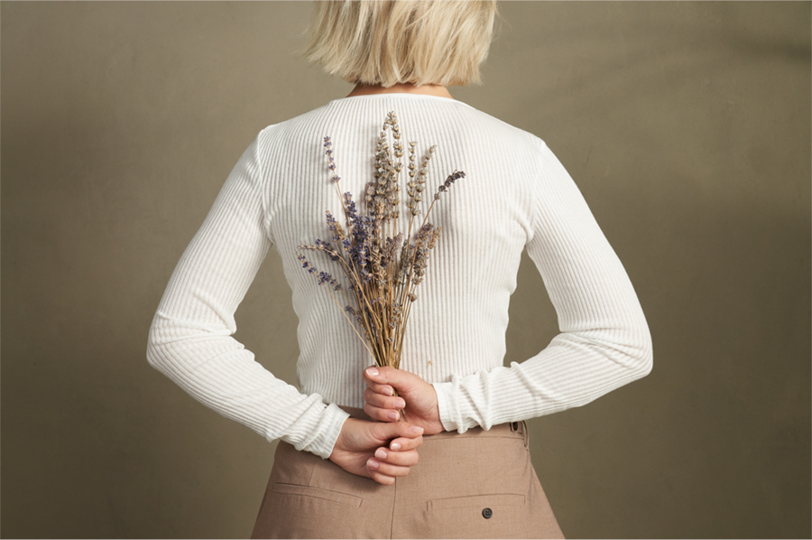 Eine Frau mit blonden schulterlangen Haaren ist von hinten zu sehen. Sie hält einen Strauß Lavendel hinter dem Rücken mit beiden Händen.